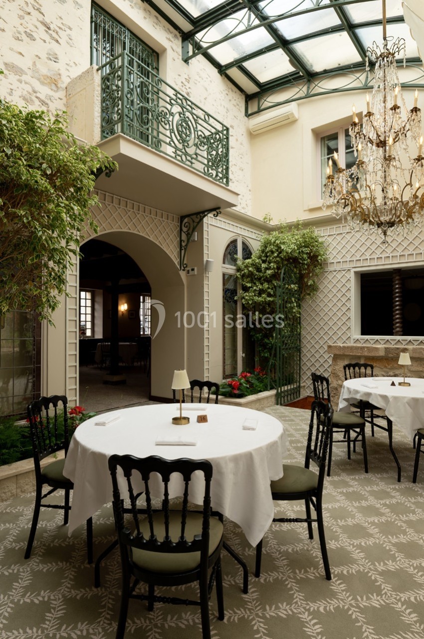 Salle lumineuse avec verrière, tables rondes dressées, chaises noires et décor végétal dans un cadre élégant.