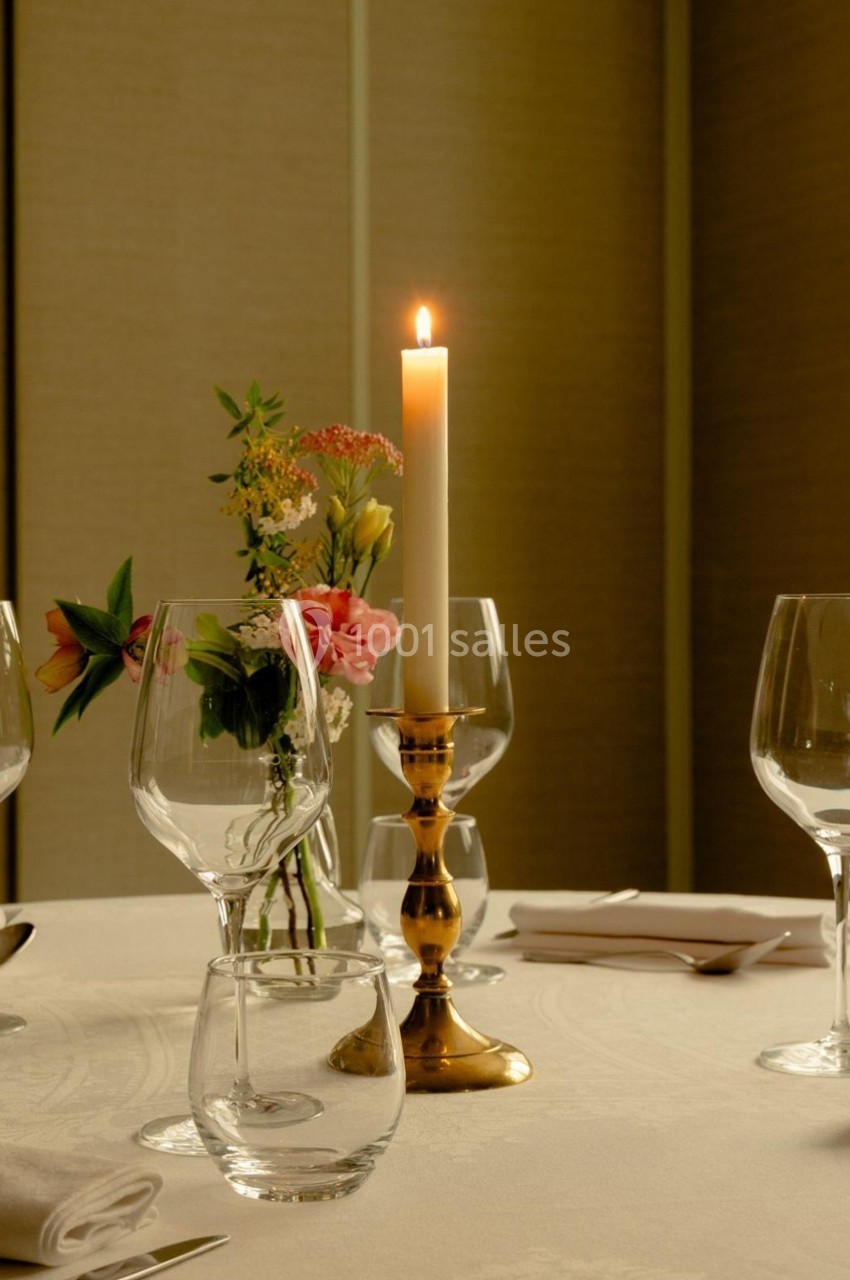 Bougie allumée sur un chandelier doré au centre d'une table dressée avec verres, fleurs et serviettes.