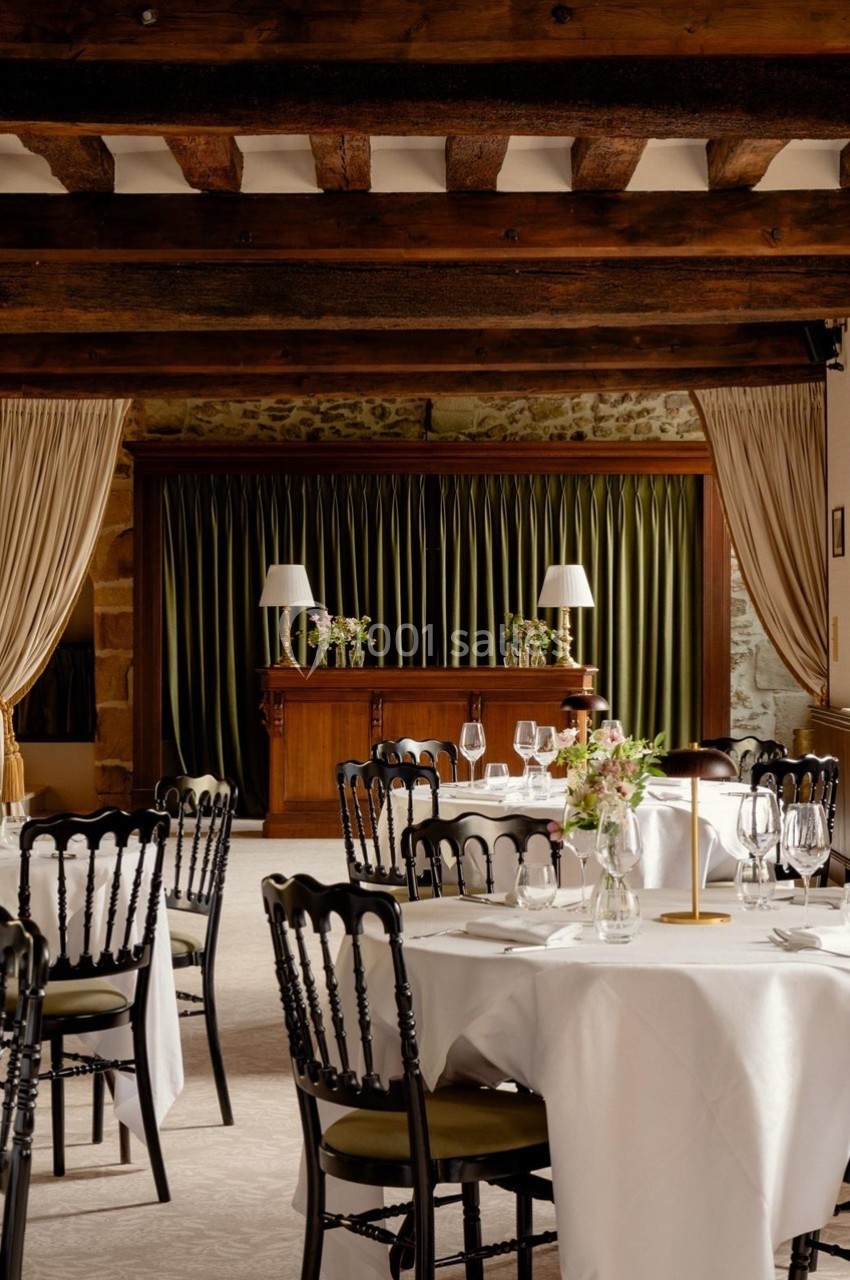 Salle de restaurant élégante avec tables dressées, chaises noires, nappes blanches et un bar en bois devant des rideaux…