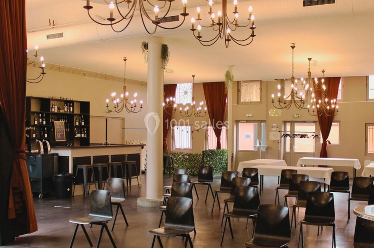 Salle lumineuse avec chaises noires, tables blanches, bar en arrière-plan et lustres suspendus.