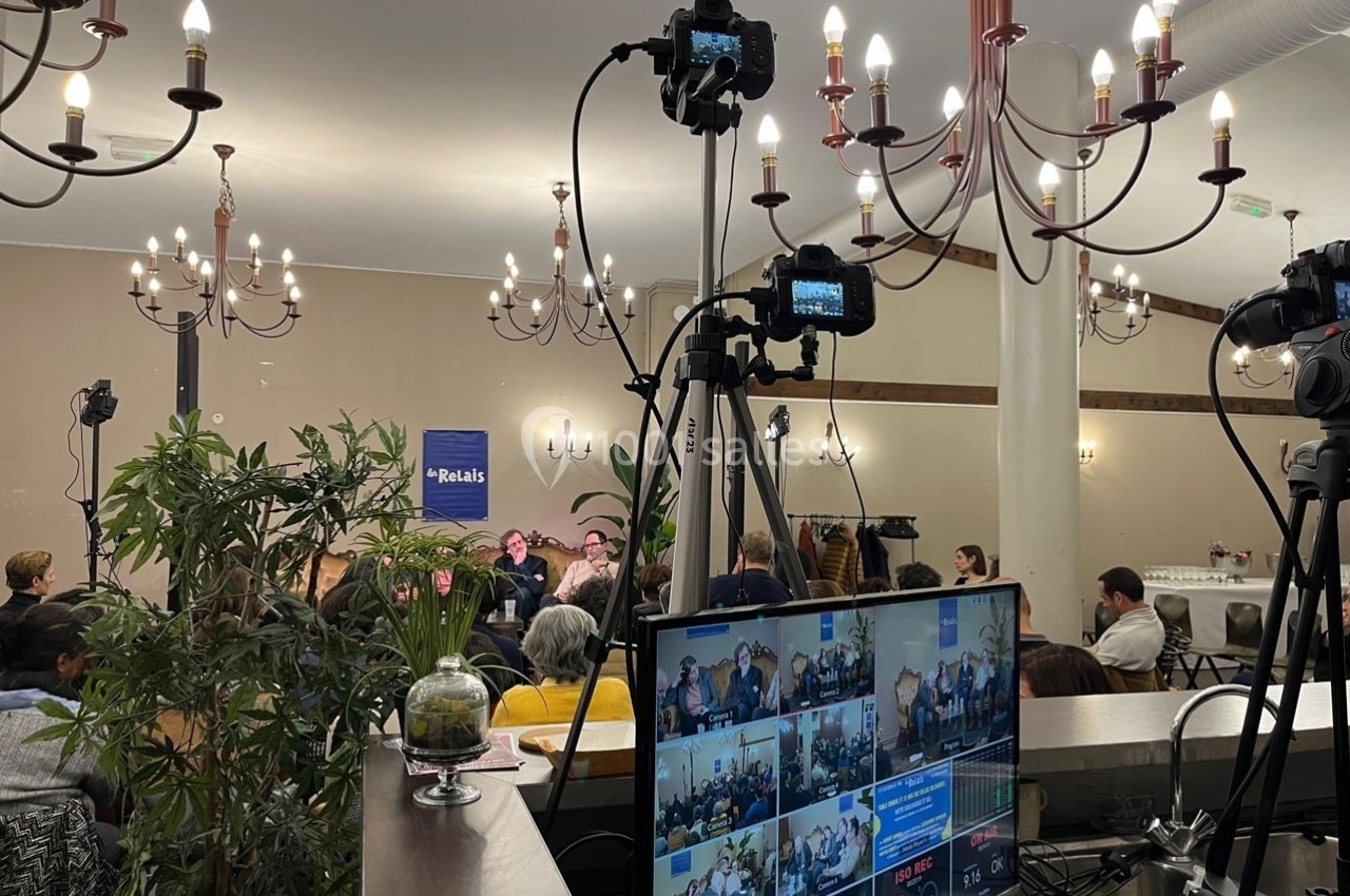 Salle de conférence avec public, caméras sur trépieds et écran affichant plusieurs vues en direct.
