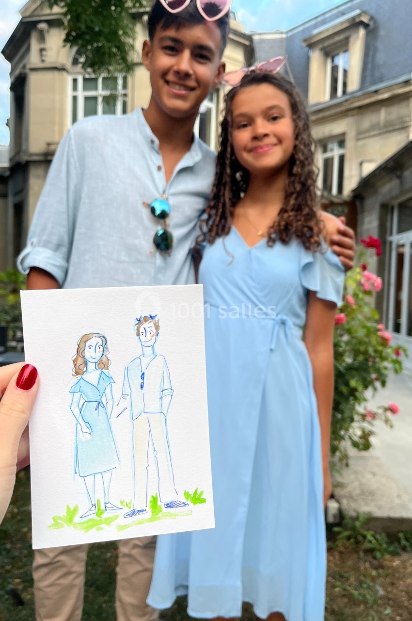 Un dessin à la main représentant un couple est tenu devant deux personnes posant dans un jardin devant un bâtiment.