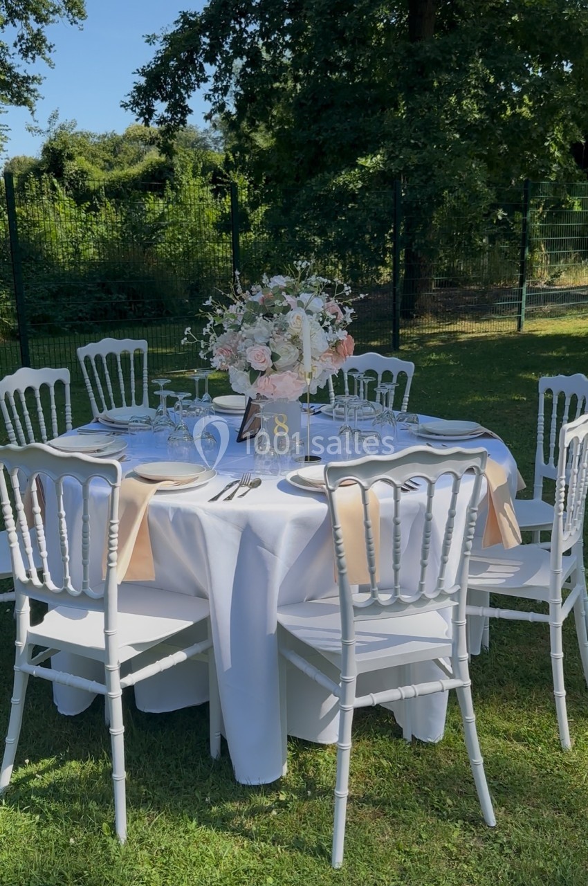 Table ronde dressée en extérieur avec nappes blanches, chaises blanches et centre de table floral.