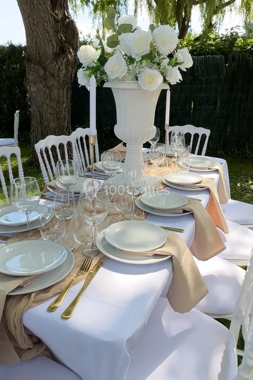 Table élégamment dressée en extérieur avec vaisselle blanche, couverts dorés et centre de table floral blanc.