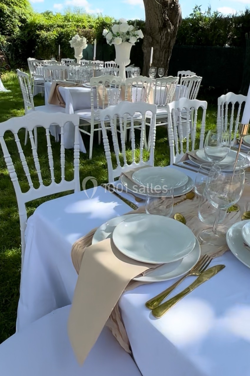 Table dressée en extérieur avec nappes blanches, vaisselle élégante, couverts dorés et chaises blanches sur pelouse.