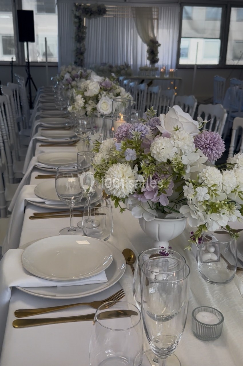 Table décorée pour un événement avec vaisselle blanche, couverts dorés et arrangements floraux blancs et violets.