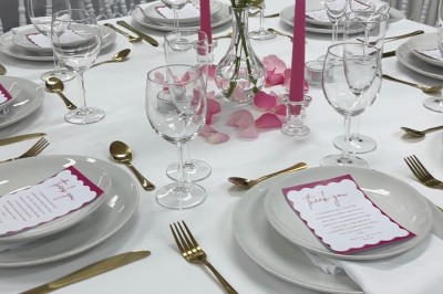 Table élégamment dressée avec nappes blanches, vaisselle raffinée, bougies roses et décoration florale assortie.