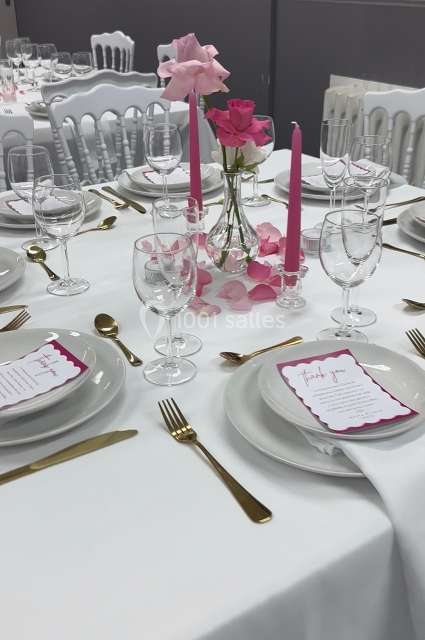 Table élégamment dressée avec nappes blanches, vaisselle dorée, menus roses et centre de table floral rose.