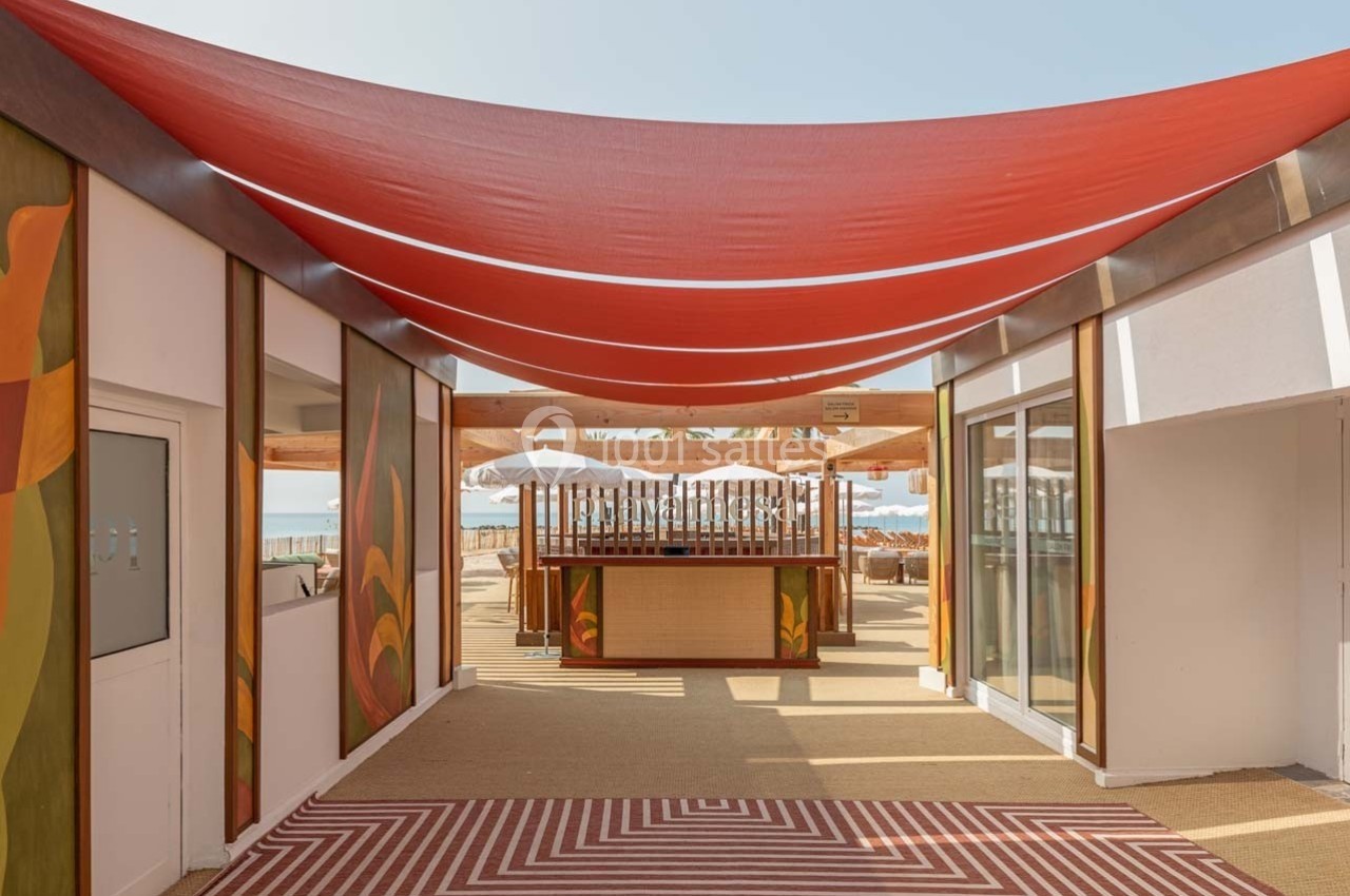 Vue d'une terrasse extérieure avec une toile rouge tendue, des motifs colorés et un bar en arrière-plan.