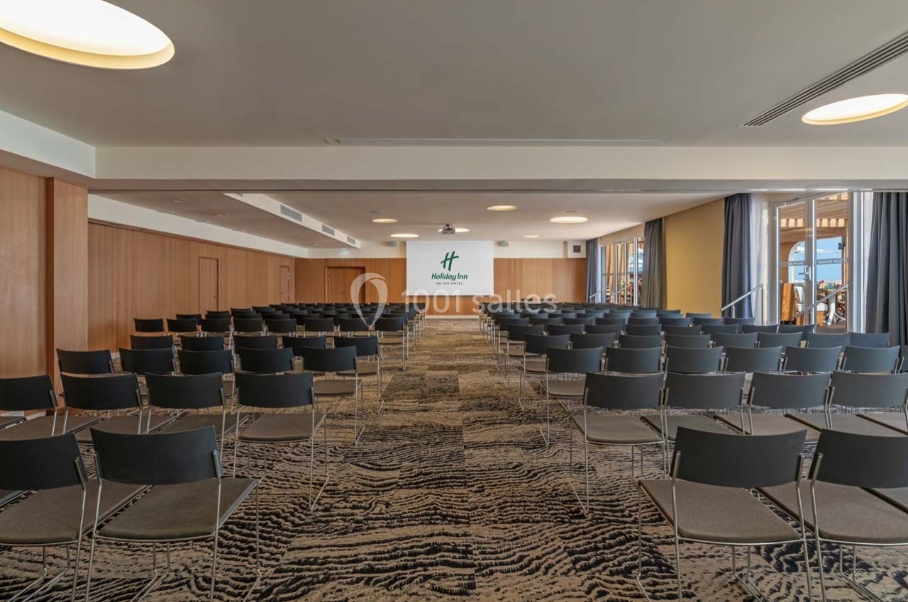 Salle de conférence avec rangées de chaises noires, écran de projection et éclairage naturel par de grandes fenêtres.