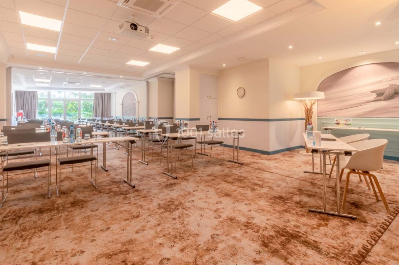 Salle de réunion lumineuse avec tables, chaises, bouteilles d'eau et équipement audiovisuel, vue sur des fenêtres.