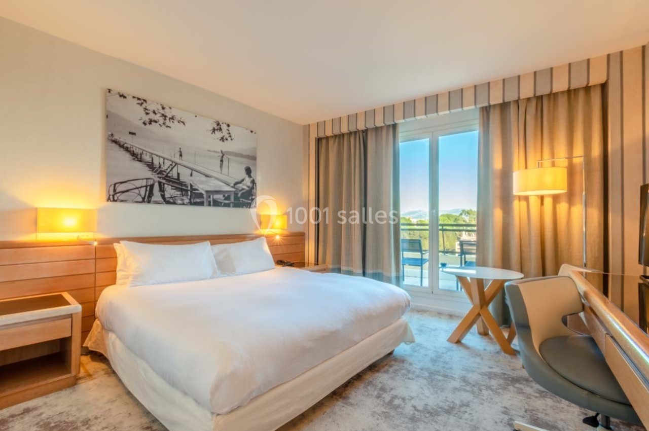 Chambre d'hôtel lumineuse avec un grand lit, un bureau, des rideaux ouverts et une vue sur un balcon et la nature.