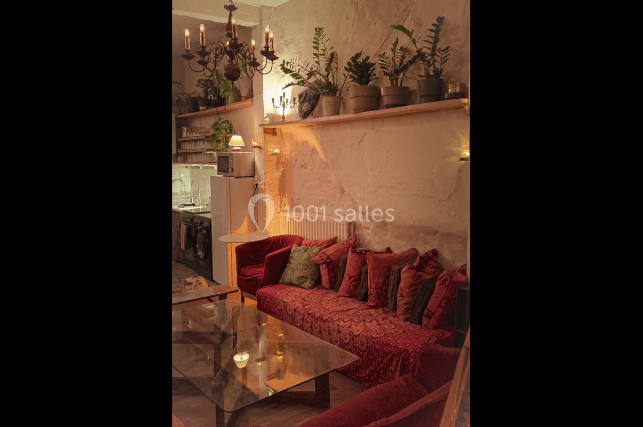 Salon chaleureux avec canapé rouge, table en verre, plantes en pot et éclairage tamisé sous un lustre ancien.