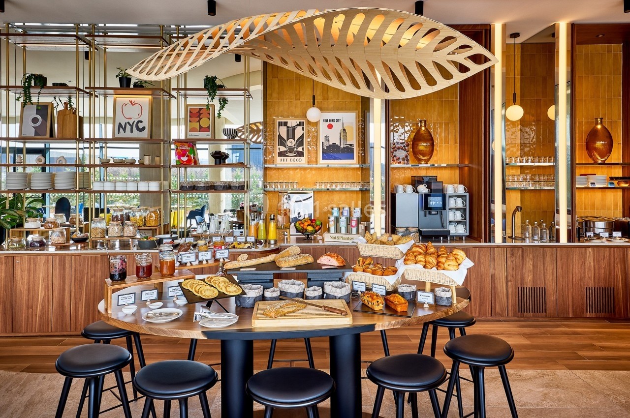 Buffet de petit-déjeuner avec viennoiseries, pains, confitures et boissons dans un espace lumineux et moderne.