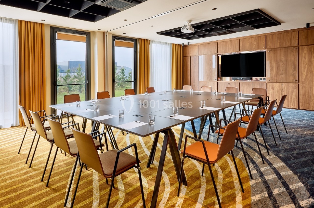 Salle de réunion lumineuse avec une grande table rectangulaire, des chaises orange et un écran mural.