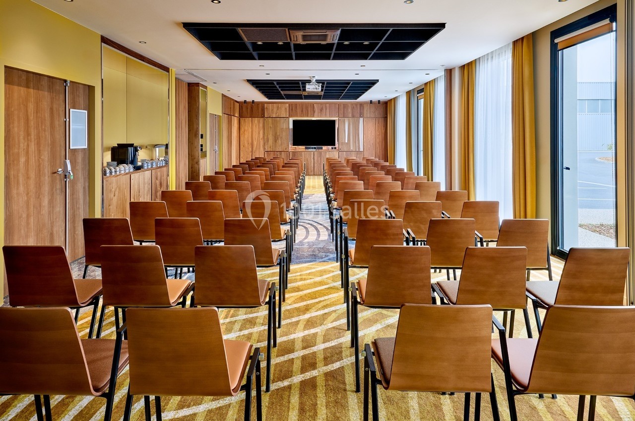 Salle de conférence lumineuse avec rangées de chaises en bois, écran de projection et grandes fenêtres.