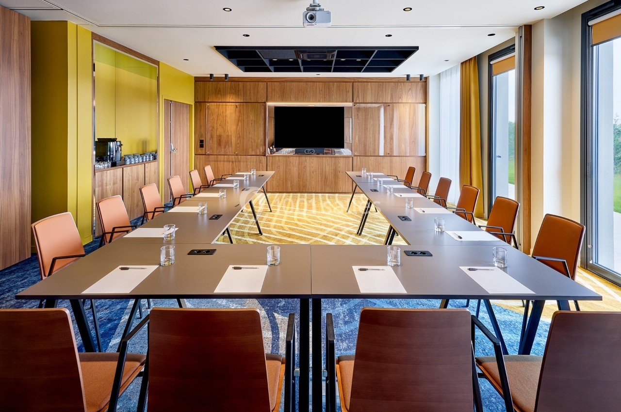 Salle de réunion lumineuse avec tables disposées en U, chaises en bois, écran mural et grandes fenêtres.