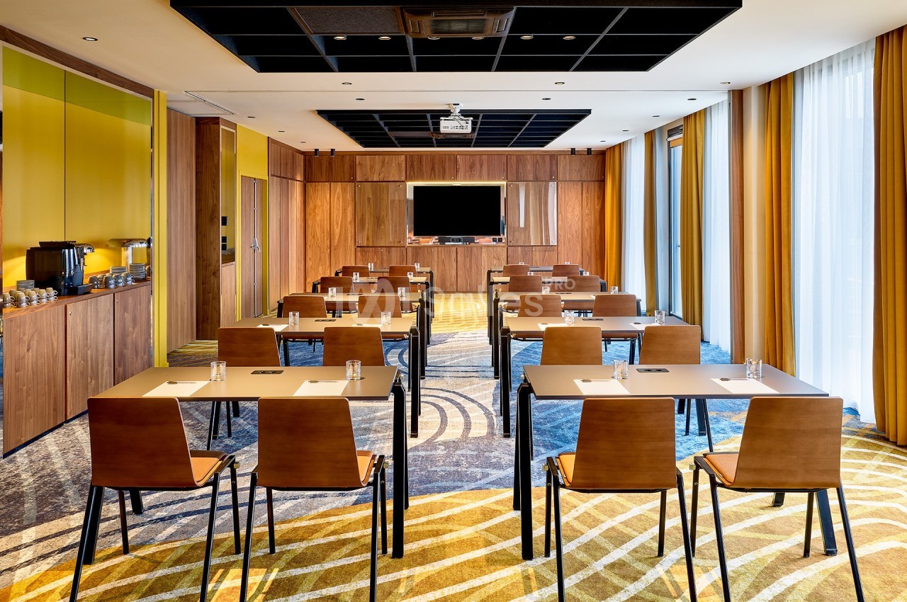 Salle de réunion lumineuse avec tables alignées, chaises en bois, écran mural et rideaux jaunes.