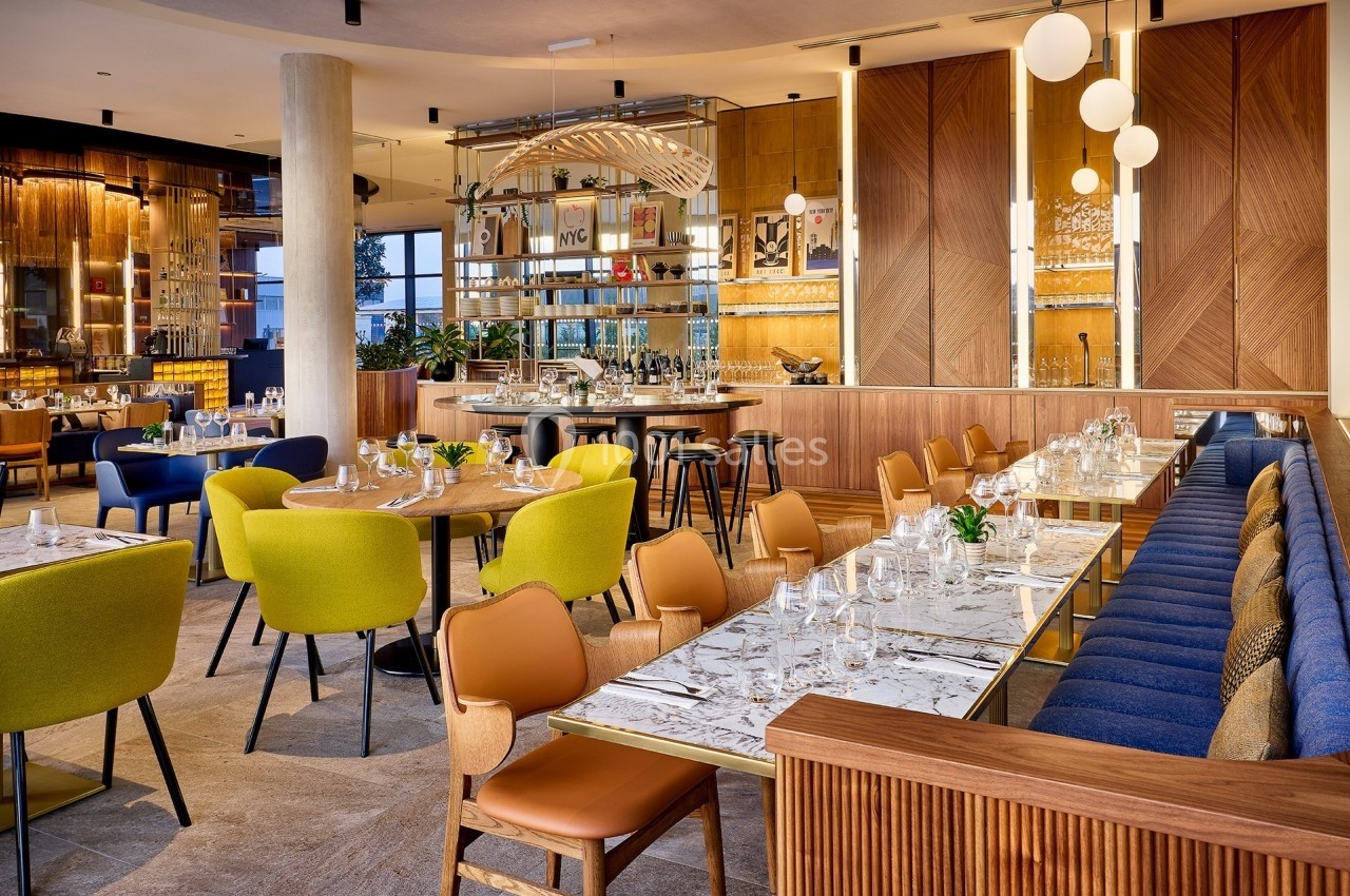 Salle de restaurant moderne avec tables dressées, chaises colorées, éclairage chaleureux et bar central élégant.