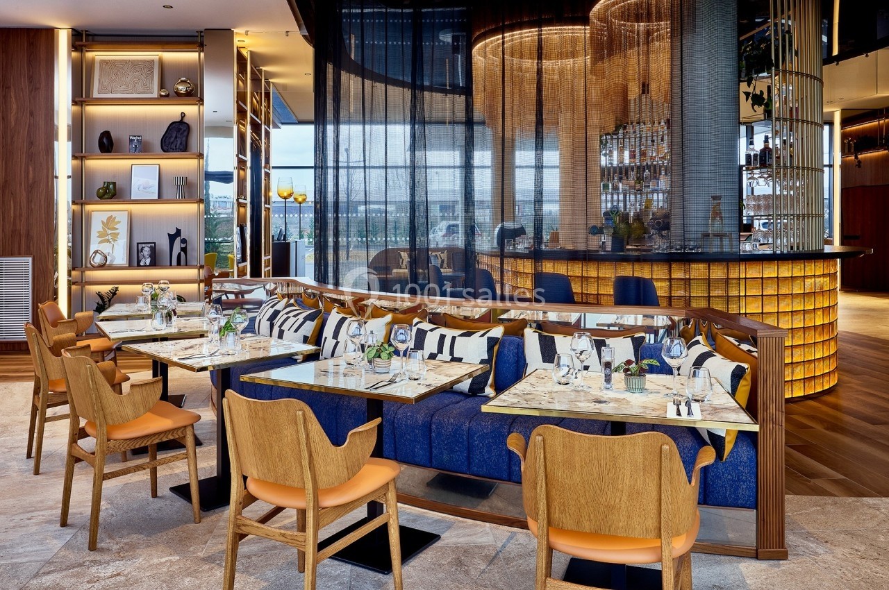 Salle de restaurant moderne avec tables dressées, banquettes bleues, chaises en bois et bar éclairé en arrière-plan.