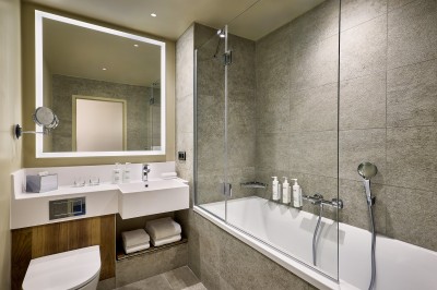 Salle de bain moderne avec baignoire, paroi vitrée, lavabo avec miroir éclairé et étagère contenant des serviettes.