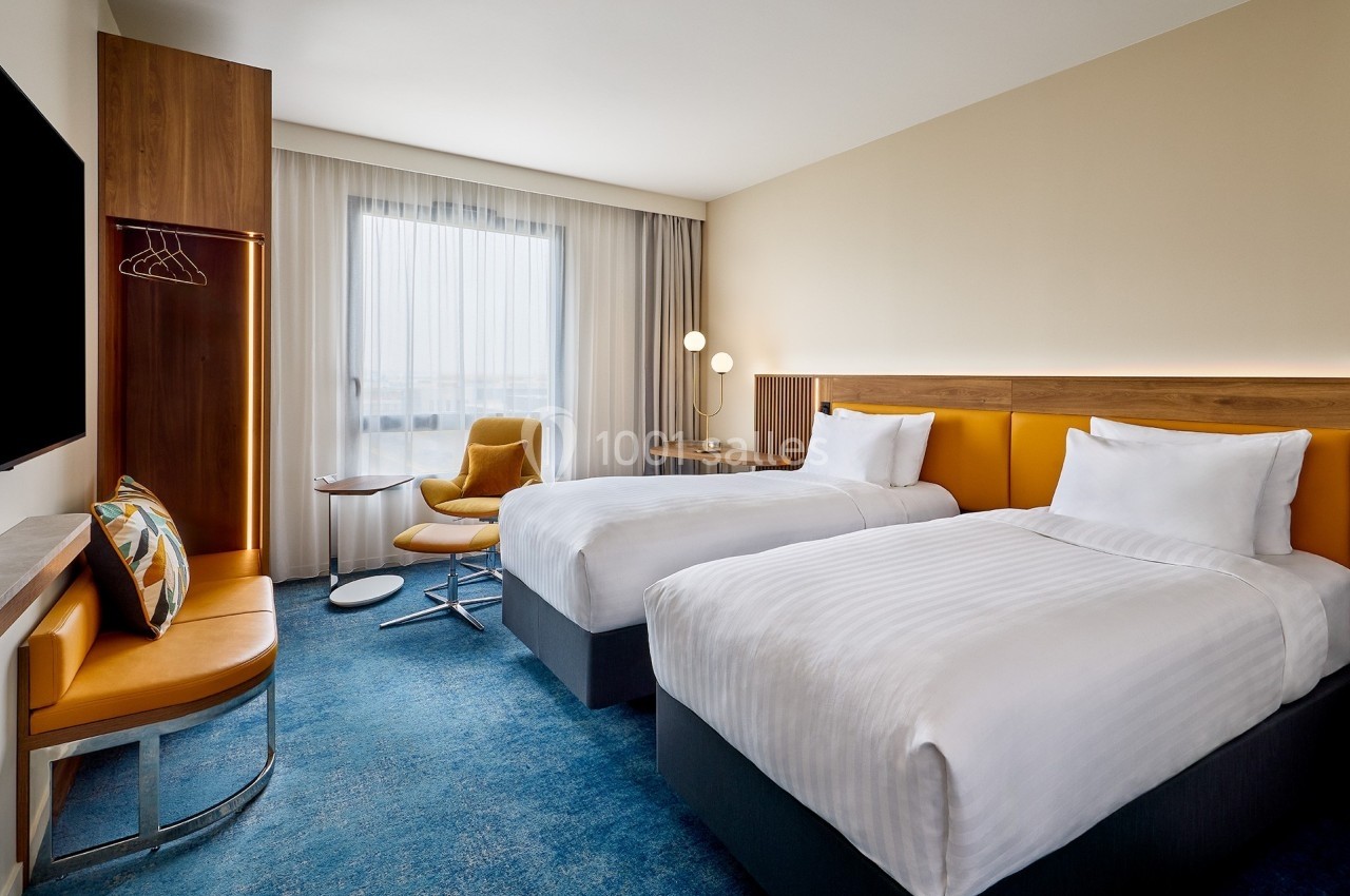 Chambre d'hôtel moderne avec deux lits simples, fauteuil, bureau, télévision murale et grande fenêtre avec rideaux.