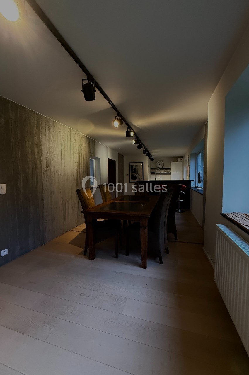 Salle à manger avec table en bois, chaises sombres, mur en bois à gauche et grande fenêtre à droite.