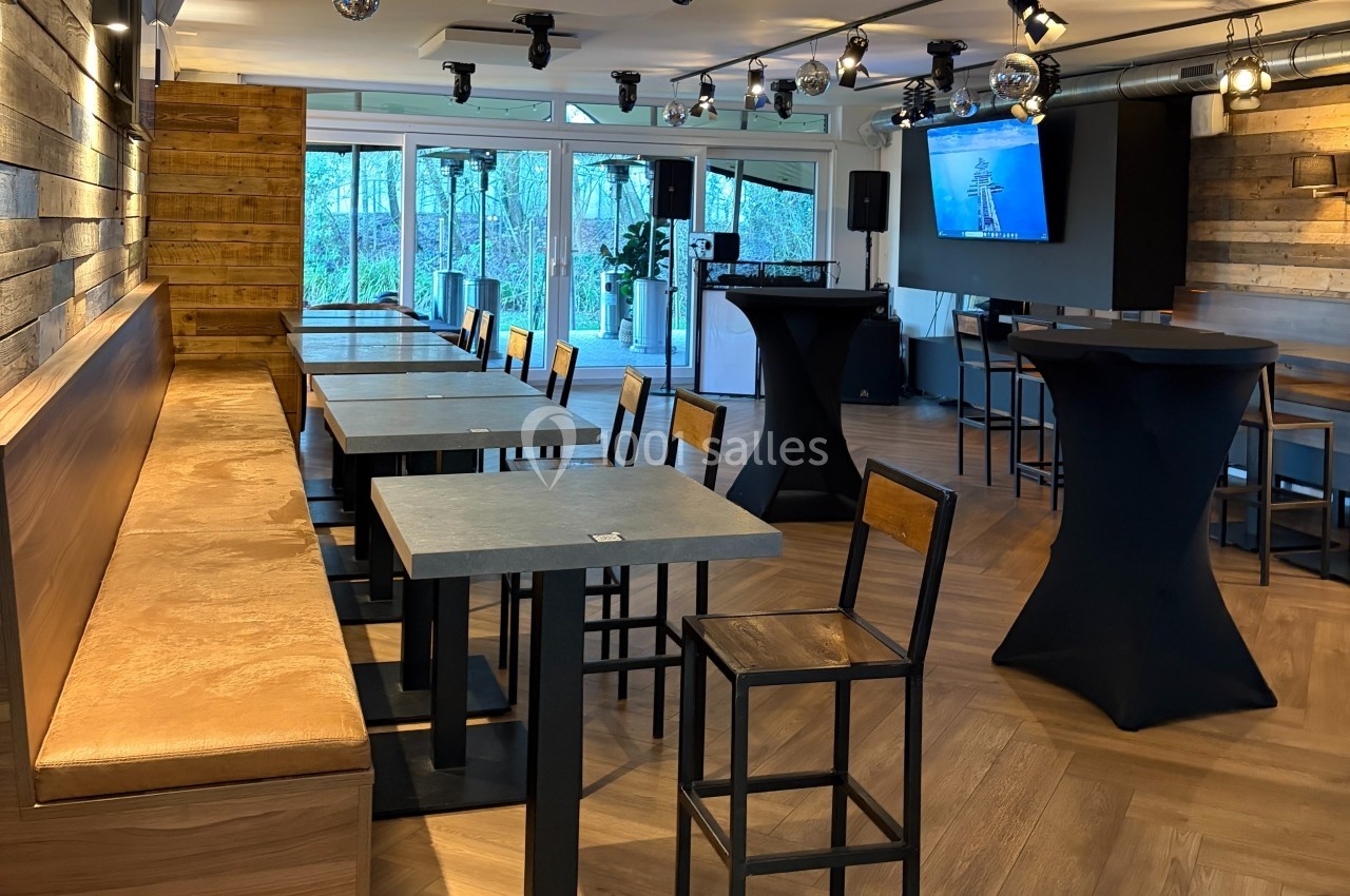 Salle lumineuse avec tables, chaises et mange-debout, décorée de bois et équipée d'un écran mural.