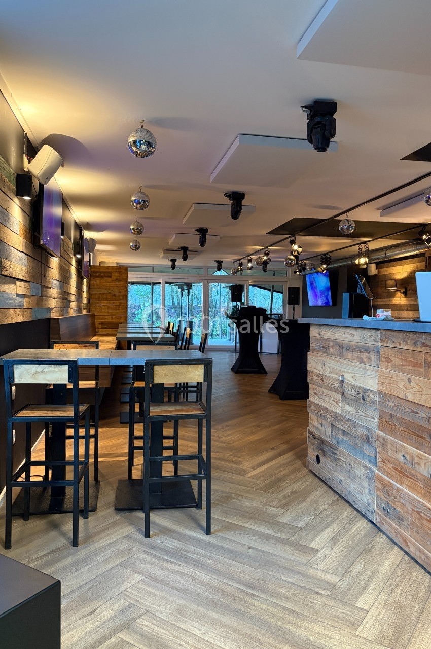 Intérieur d'un bar moderne avec mobilier en bois, parquet clair, tables hautes et écrans accrochés aux murs.