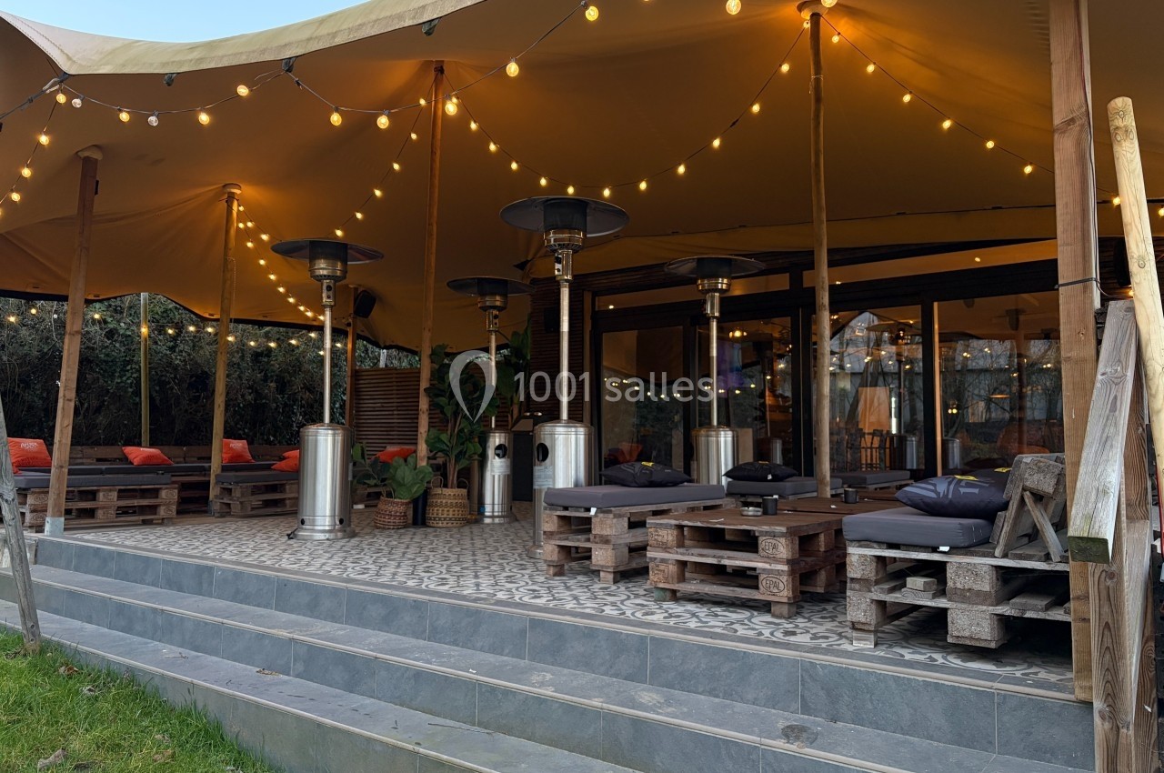 Terrasse couverte avec éclairage suspendu, mobilier en palettes, chauffages extérieurs et coussins sur les assises.