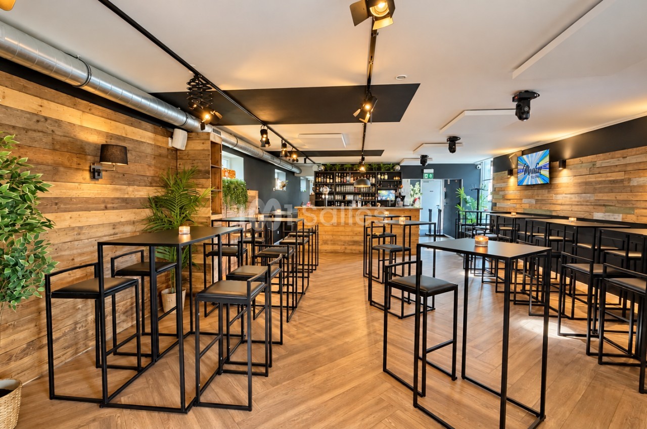 Salle de réception aux murs en bois clair, avec tables hautes noires entourées de tabourets, bar en fond et éclairage sur rails au plafond.