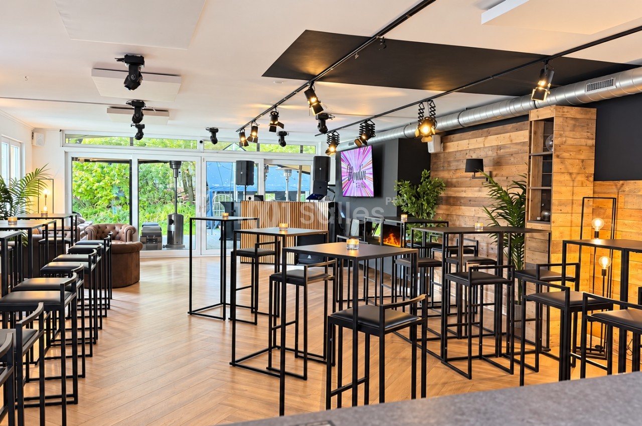 Salle de réception lumineuse aménagée en espace debout avec hauts tables et tabourets noirs, mur en bois, spots sur rail et baies vitrées donnant sur un jardin.