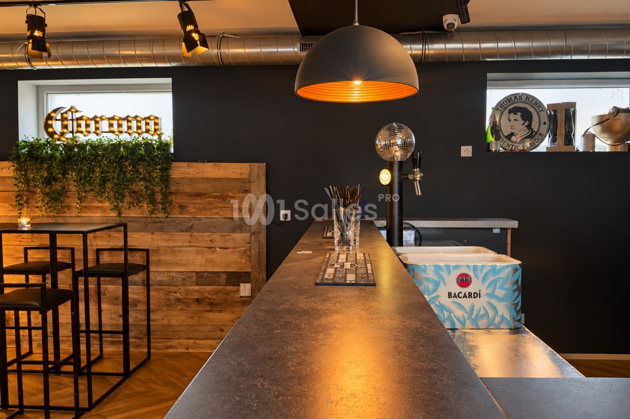 Comptoir de bar en bois sombre éclairé par une suspension noire, avec tabourets hauts en métal, étagère murale et seaux à bouteilles sur le plan de travail. Let me check length: roughly 165 characters, too long. Need ≤125. Comptoir de bar sombre sous une