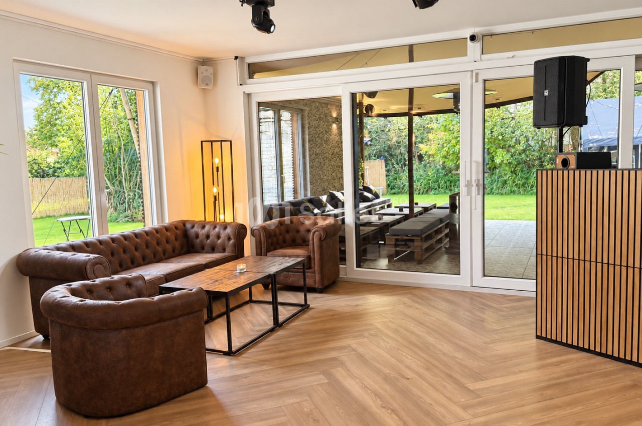 Salon aménagé avec canapés Chesterfield en cuir brun, tables basses en bois et métal, et enceinte sur pied en bois face à de grandes baies vitrées donnant sur un jardin.