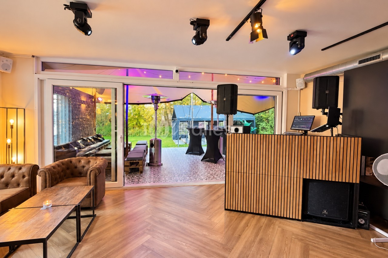 Salle lounge au parquet clair avec canapés en cuir capitonné, bar DJ en bois et enceintes, ouverte sur une terrasse sous tente avec tables hautes.