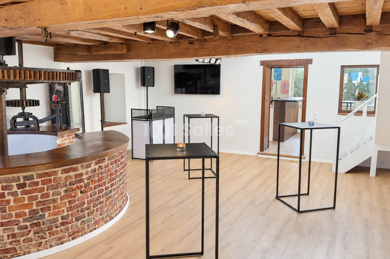 Salle aux poutres apparentes avec bar courbe en briques et ancien pressoir en bois, plusieurs tables hautes noires et enceintes sur pied.