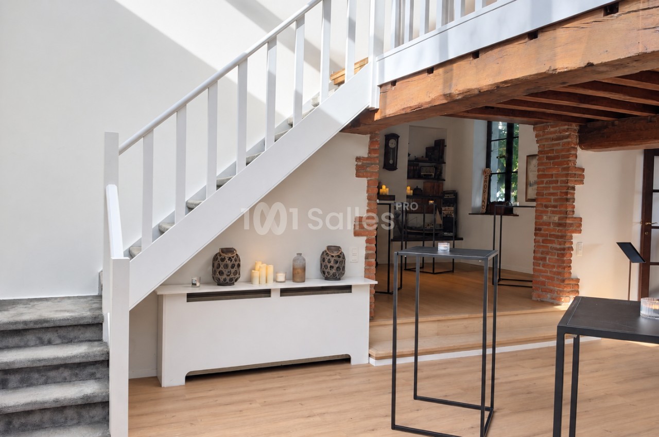 Escalier blanc à rampe en bois menant à une mezzanine, poutres apparentes et murs en briques dans une pièce claire aux sols en parquet clair.