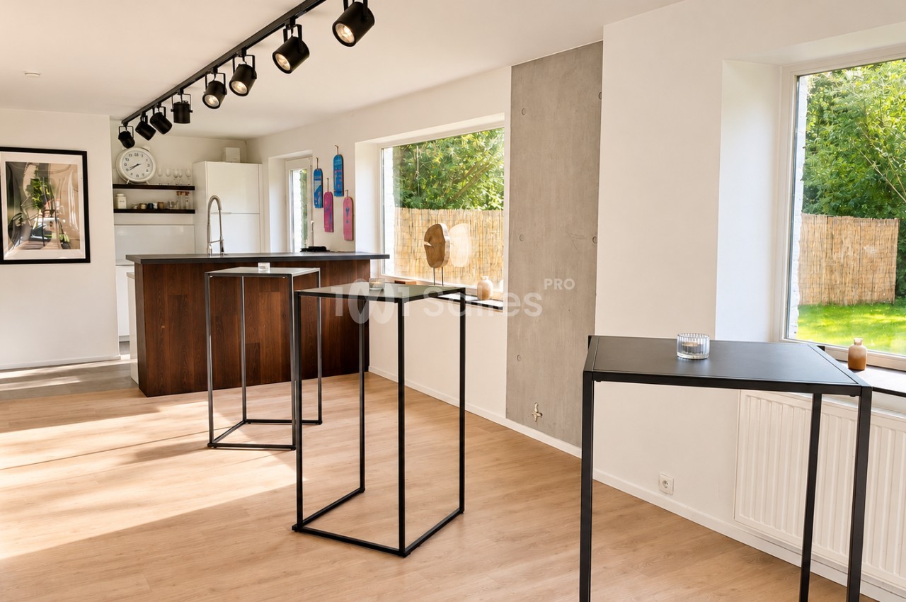 Pièce lumineuse au sol en parquet clair, avec des tables hautes noires à structure métallique, un îlot de cuisine en bois foncé et une grande fenêtre donnant sur un jardin.