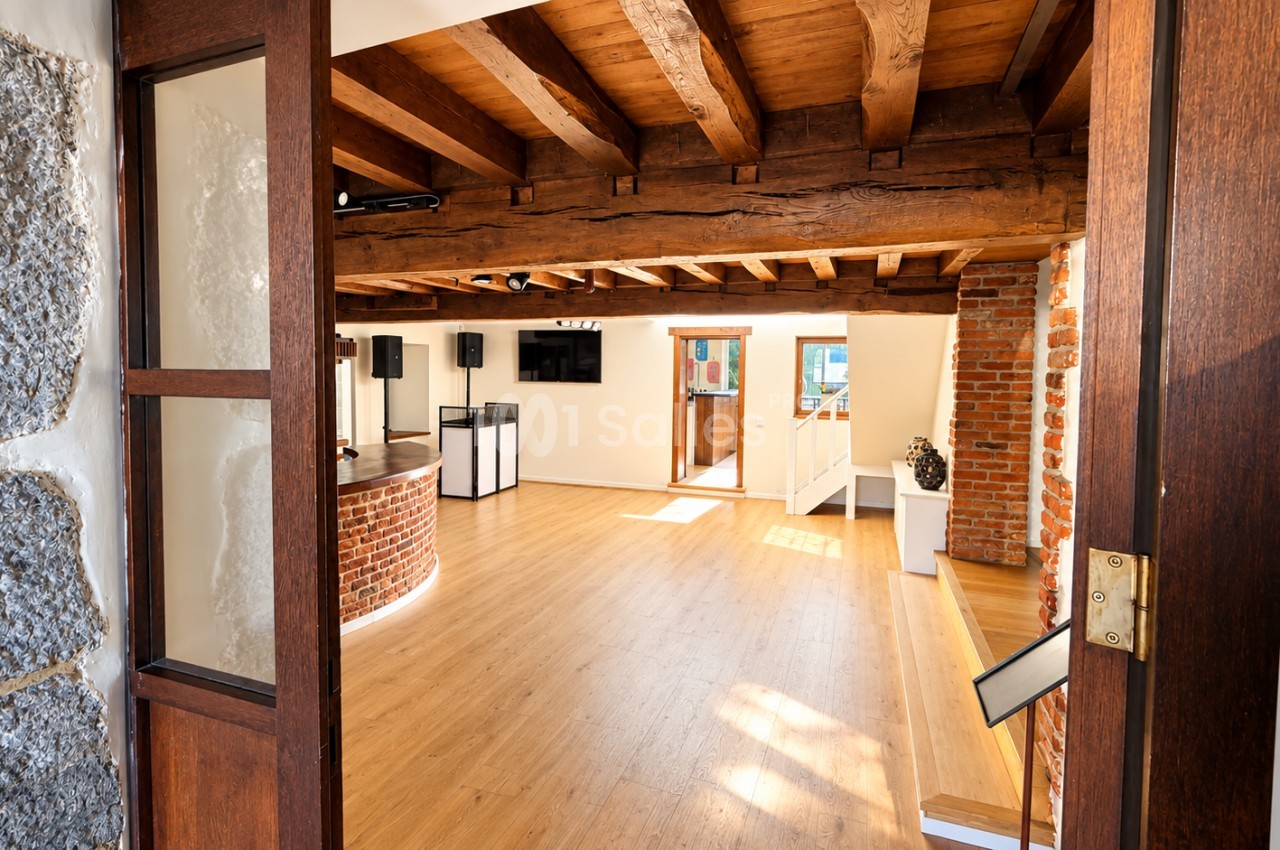 Salle vide au parquet clair sous des poutres en bois apparentes, avec piliers et murs en briques, vue depuis une porte en bois vitrée.