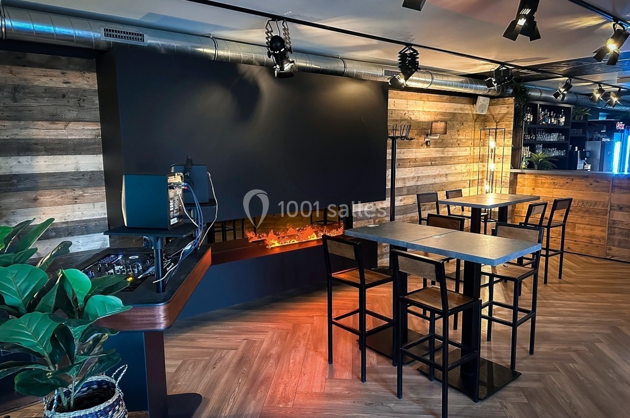 Salle moderne avec tables hautes, cheminée électrique allumée, éclairage suspendu et murs en bois.