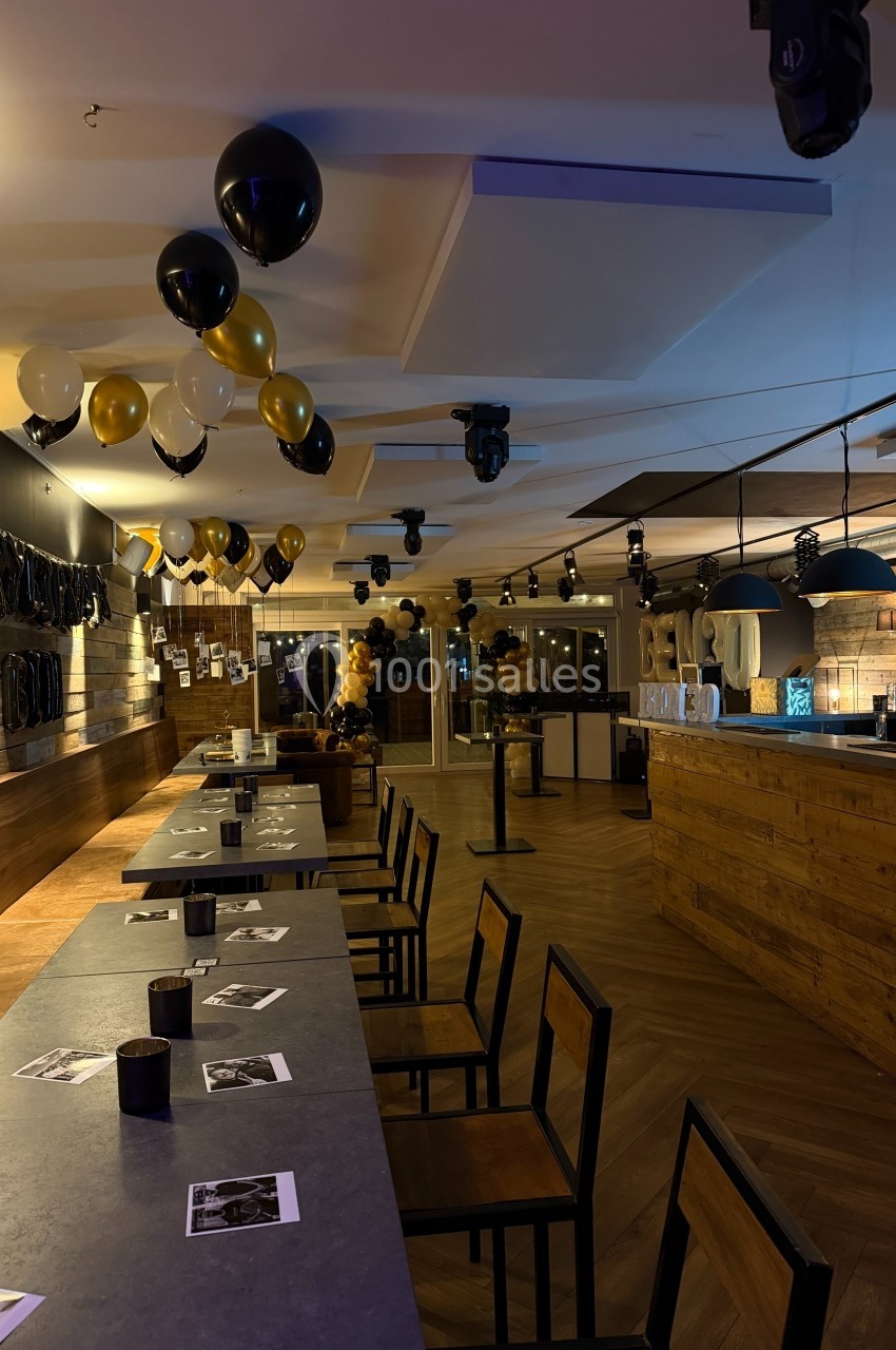 Salle décorée avec des ballons noirs et dorés, tables alignées, éclairage tamisé et bar en arrière-plan.