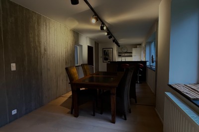 Salle lumineuse avec parquet clair, table et chaises au centre, escalier blanc, machine à coudre ancienne et tableau au mur.