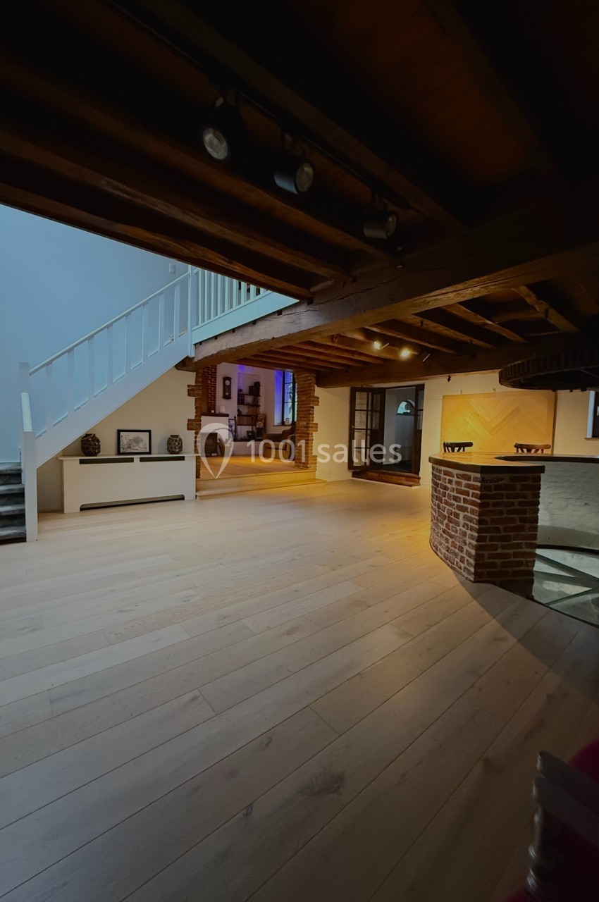 Intérieur d'une maison avec escalier blanc, poutres apparentes, parquet clair et bar en briques.