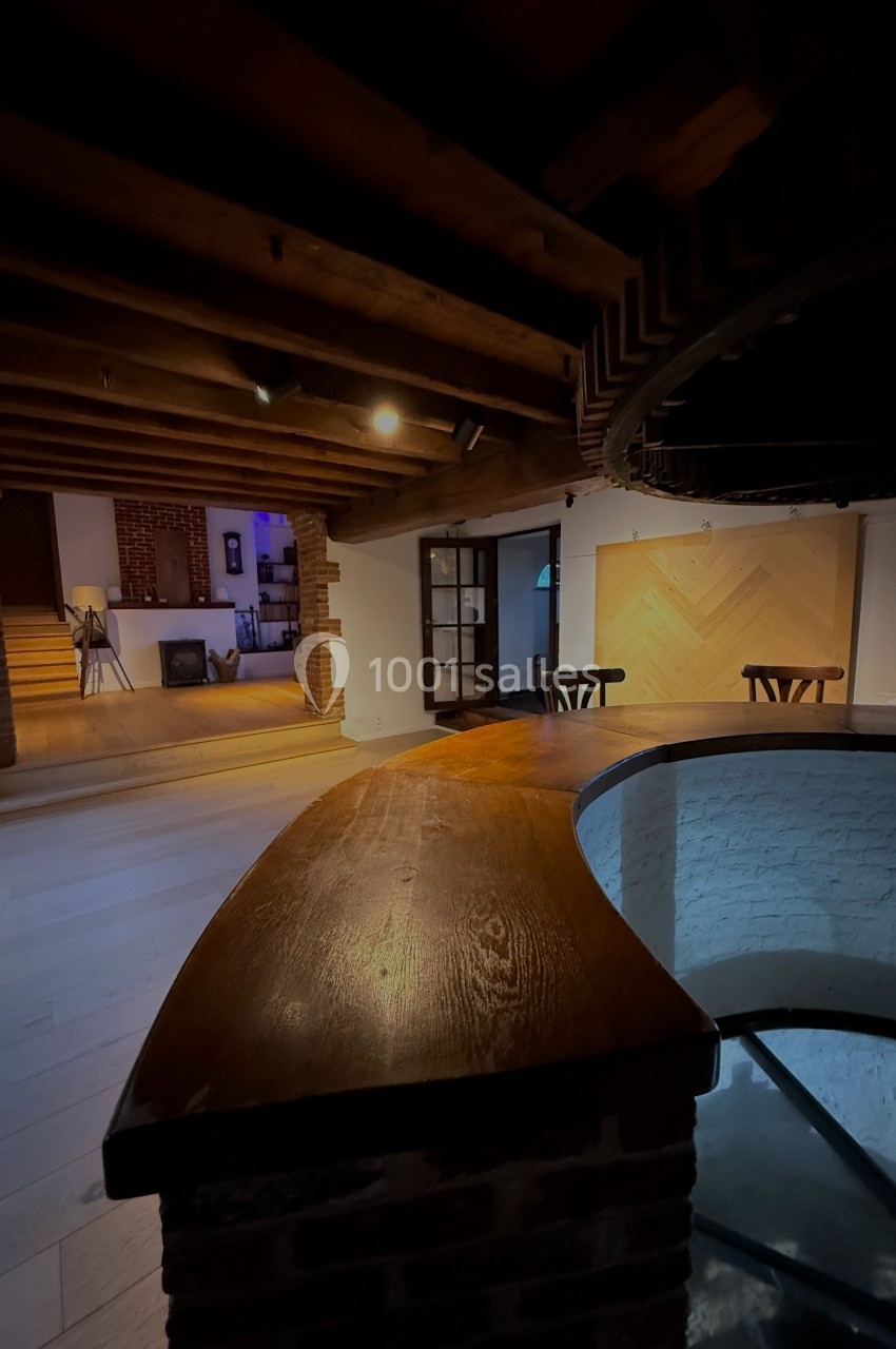 Vue intérieure d'une pièce avec plafond en poutres apparentes, escalier descendant et mobilier en bois.