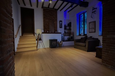 Salle lumineuse avec parquet clair, table et chaises au centre, escalier blanc, machine à coudre ancienne et tableau au mur.