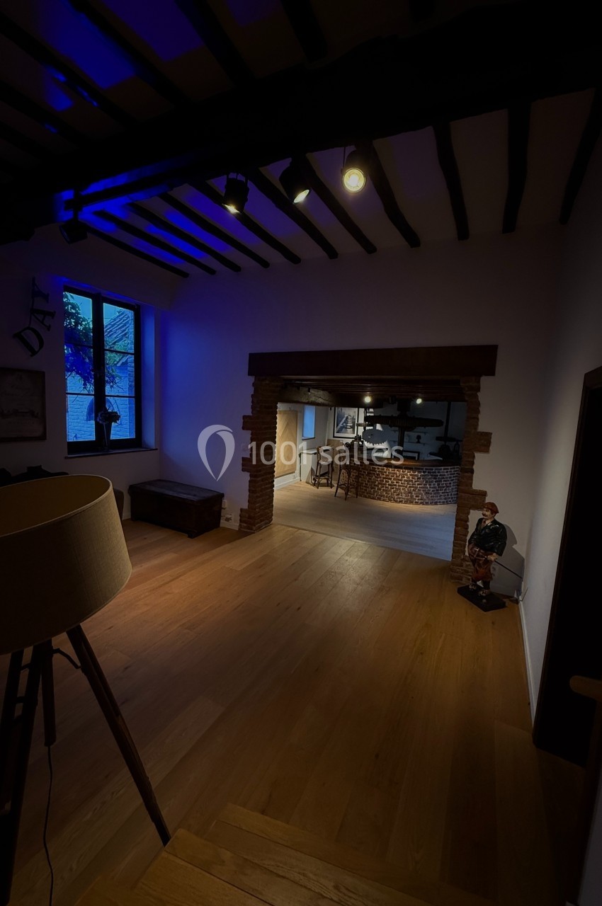 Vue d'un intérieur avec parquet en bois, poutres apparentes, éclairage tamisé et ouverture vers une cuisine.