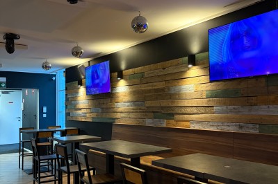 Salle lumineuse avec parquet clair, table et chaises au centre, escalier blanc, machine à coudre ancienne et tableau au mur.