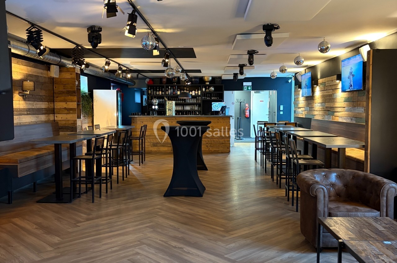 Salle moderne avec parquet en chevrons, tables hautes, bar en bois au fond et éclairage suspendu.