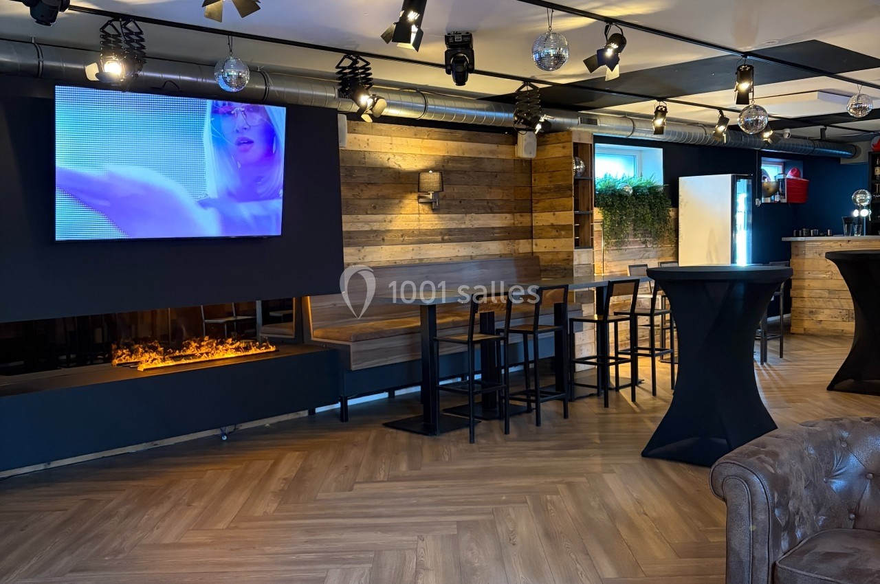 Intérieur d'un bar moderne avec écran mural, cheminée, tables hautes et décoration en bois et métal.