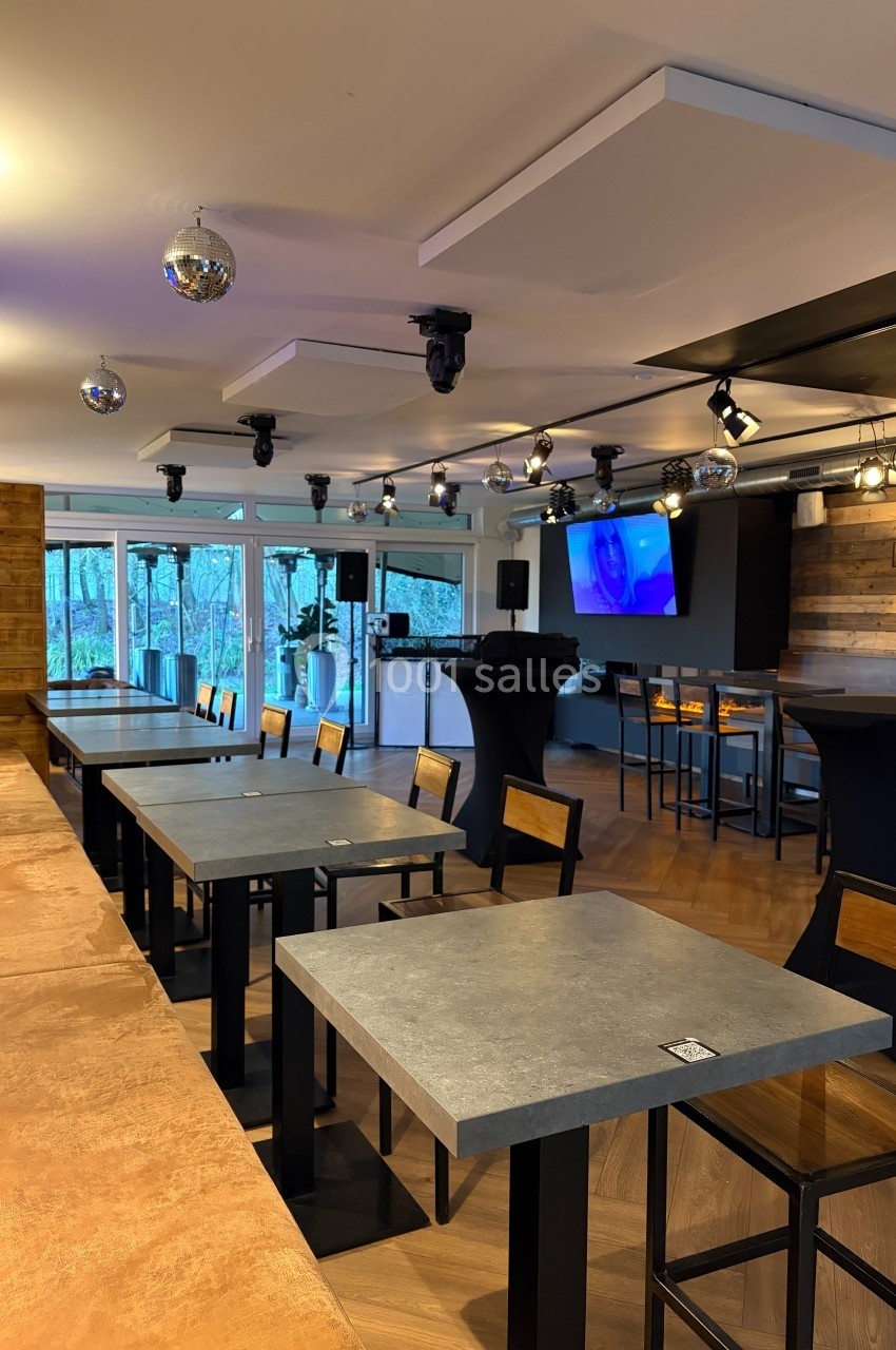 Salle moderne avec tables en béton, chaises en bois, éclairage suspendu et baies vitrées donnant sur l'extérieur.